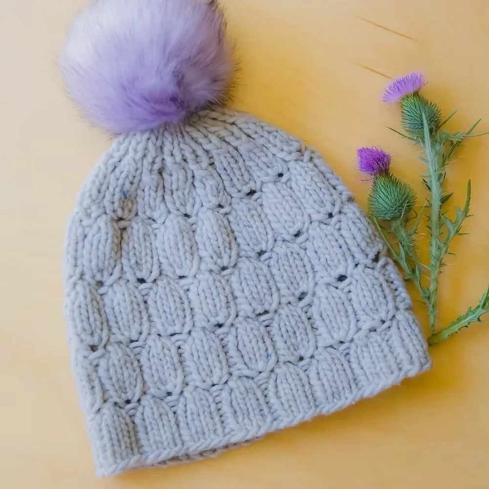 A grey knitted toque with a purple pompom lays flat on a yellow background next to a thistle plant.
