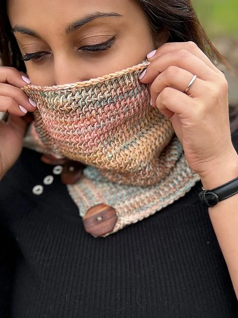 A close up of a woman wearing a cowl made with a variety of pastel colours.  The cowl is partially covering her face, from the nose down.