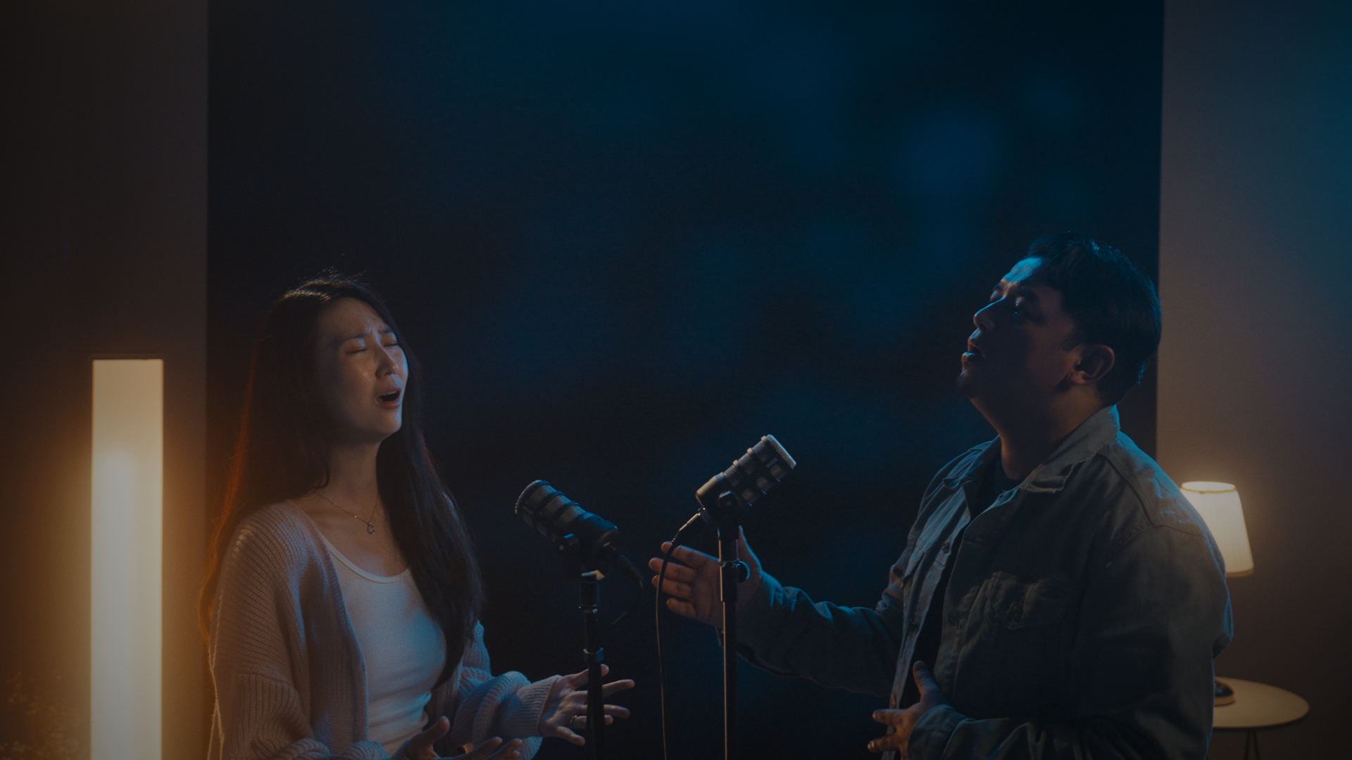 Two people singing passionately into microphones in a dimly lit recording studio.
