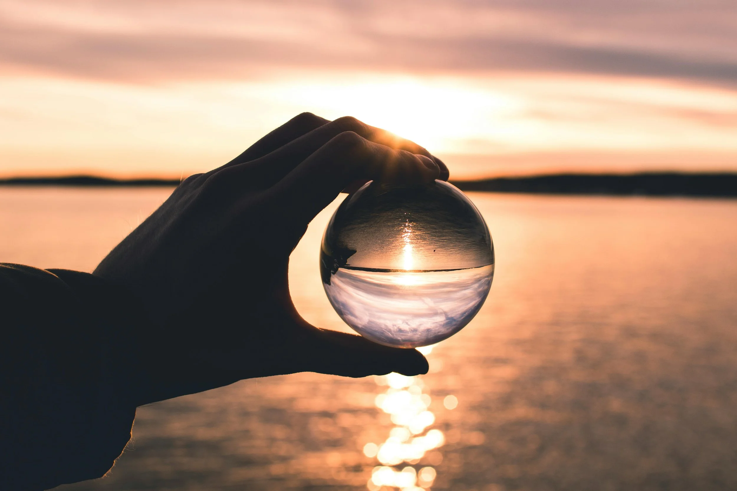 Hand holding a glass sphere up to a sunset scene, which shows upside down in the ball