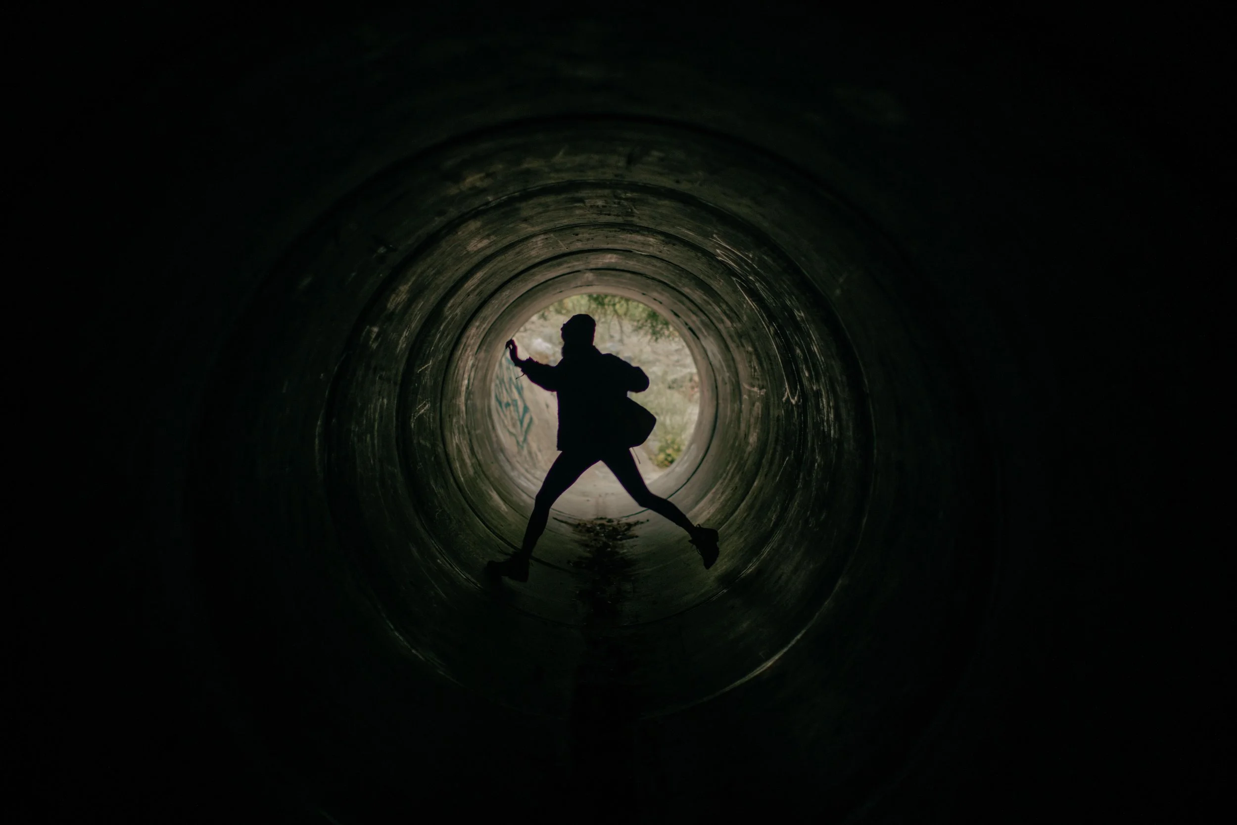 Silhouette of a person mid-stride inside a dark concrete tunnel, legs wide apart, walking towards a circle of daylight and greenery at the far end.