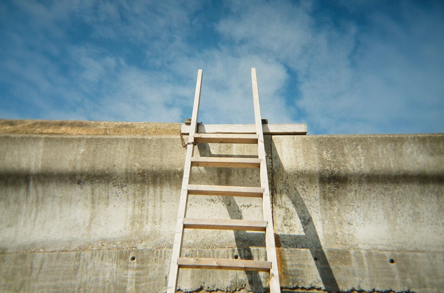 A weathered wooden ladder leans against a tall concrete wall, its top rungs reaching into a bright blue sky with scattered clouds