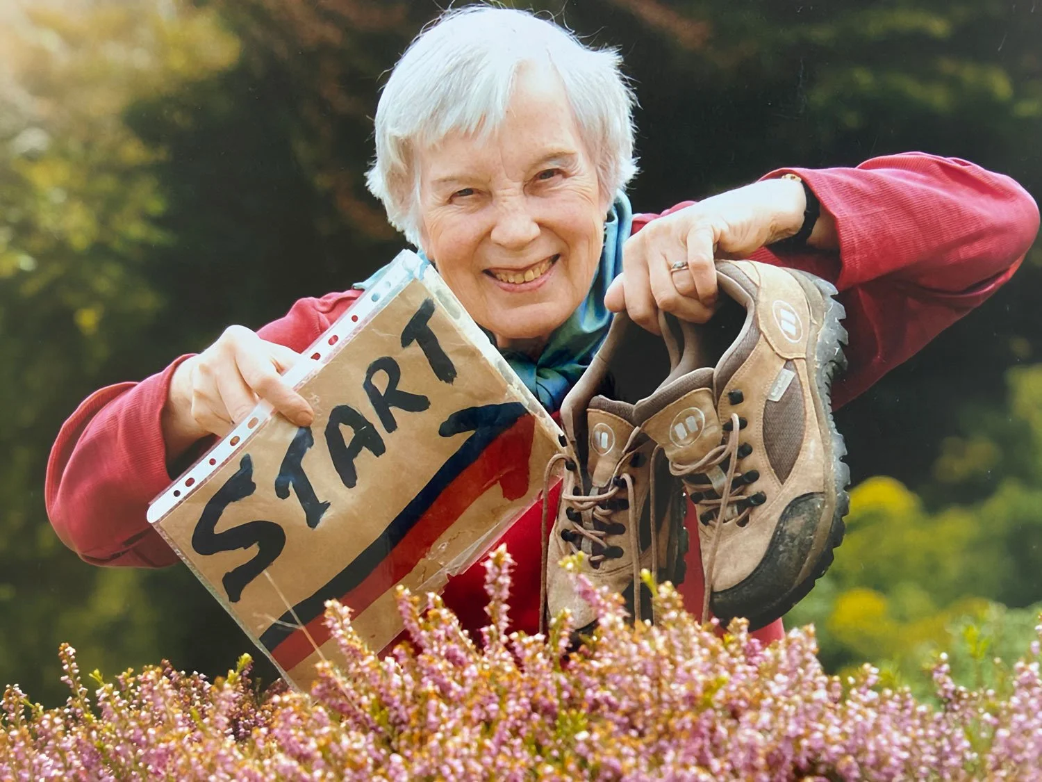 An older woman with short white hair, smiling broadly and holding a handmade cardboard 'START' sign alongside a pair of walking boots, standing in front of flowering heather.