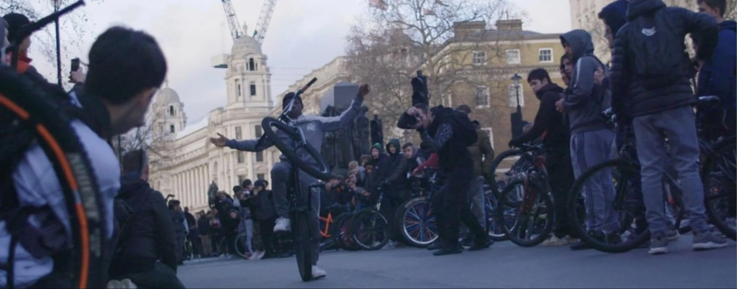Young person performing a bike trick mid-air in front of crowd of BikeStormz participants gathered in central London street with historic buildings in background
