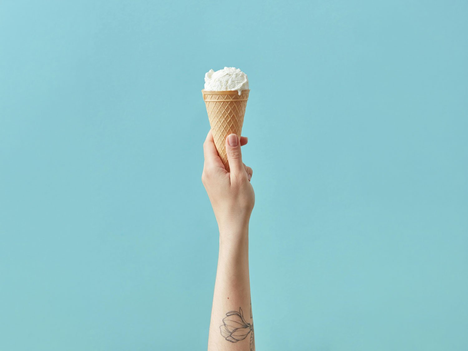 A hand holding a vanilla ice cream cone aloft against a pale blue background, with a floral tattoo visible on the wrist.