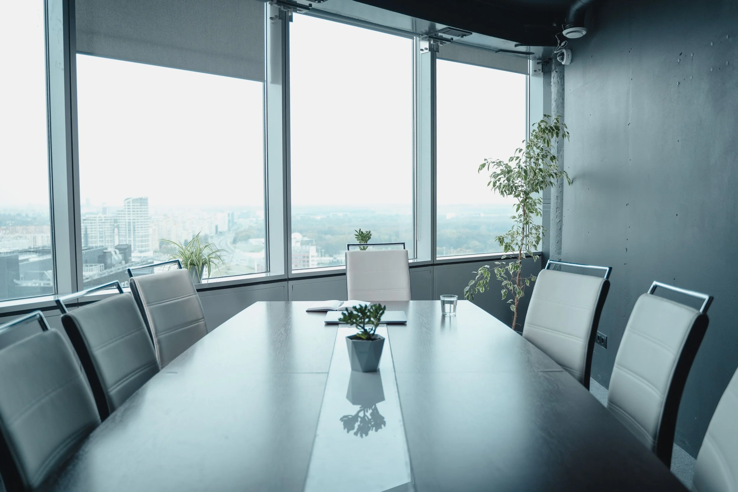 An empty boardroom with a long grey table, high-backed white leather chairs, and large windows looking out over a city skyline. A notebook and a glass of water sit at the head of the table.
