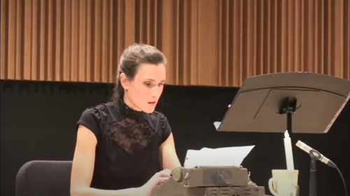 Ellen Weiser as Justine in Justine et Les Machines.Alt text: A women in a high-neck short sleeve blouse sits at a typewriter.