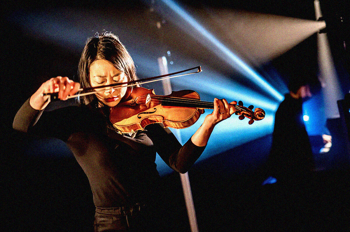 Photo Credit | Dahlia KatzAlt text: On the left, a woman in all black plays the violin with a pianist on the right hand side. Cool and warm stage lights are behind them.