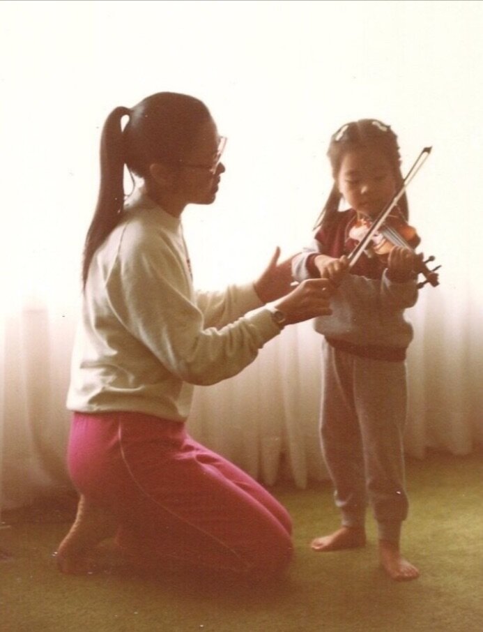 Photo of young Leslie and her mother | courtesy of artistAlt text: A woman in pink pants and a white turtleneck is crouched on the floor helping a young girl hold her violin.