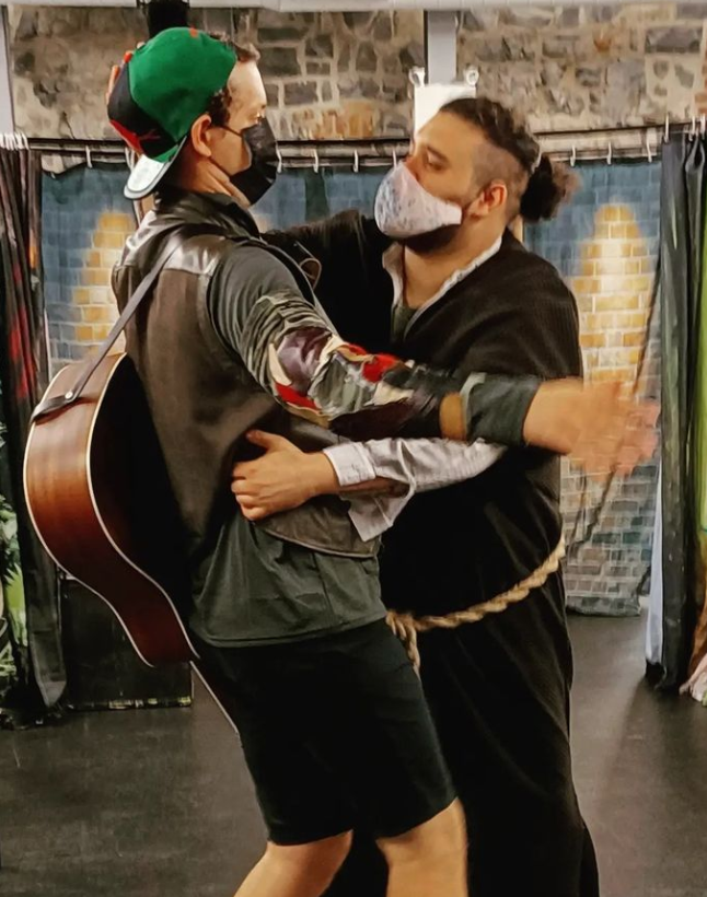 Image taken from @thelakesideplayers Instagram page. Originally captioned: “Friar Tuck saves Robin from the Vampire Squids.” Andrew Cameron playing Robin Hood on the left, Kareem Vaude as Friar Tuck on the right. Costume design by Rachel Forbes. Alt text: Vaude holds Cameron in a strong grip, their gaze intense. Friar wears a long brown robe with a rope tied as a belt, while Robin wears a brown vest over a green shirt and black shorts, with a green hat and guitar on his back. The background is the brick-walled Tett Centre with a brick curtain backdrop in front of it.