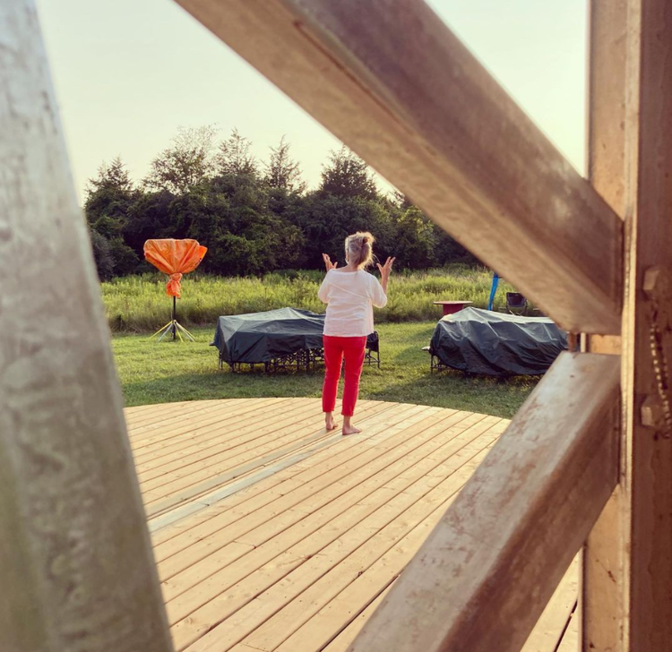 Rehearsal photo taken from the @flightfestivalpec Instagram page. Captioned: “Carol Anderson rehearsing her new solo “Owl” on the @festivalplayers gorgeous outdoor stage. The solo addresses the pain of stolen children from the perspective of a watching owl. Devastating and beautiful. @flightfestivalpec #danceisforeveryone @countyarts @eddiepec”.Alt text: A woman with grey hair wearing red trousers and a white shirt has her back turned to the camera, standing on a wooden stage. Two tarped shapes sit before an expanse of grasses, trees and sky.