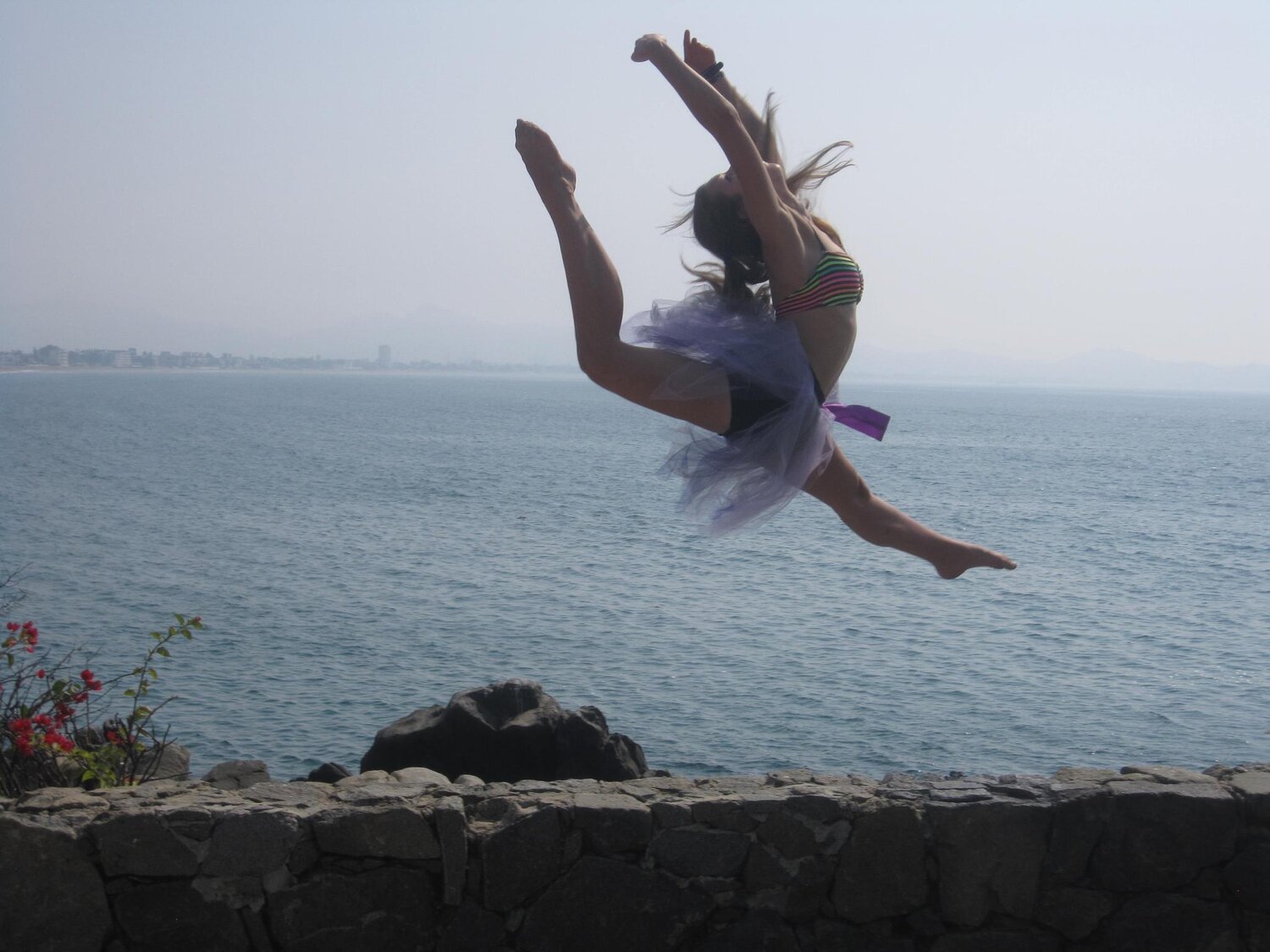 Promotional photo taken from the Festival Players of Prince Edward County’ Facebook page. Captioned: “FLIGHT: A festival of dance in its natural habitat between earth and sky, where communities connect.”Alt text: A dancer wearing a striped top and blue tutu is softly silhouetted by a bright expanse of water. The dancer is jumping, with legs extended outwards and arms upwards.