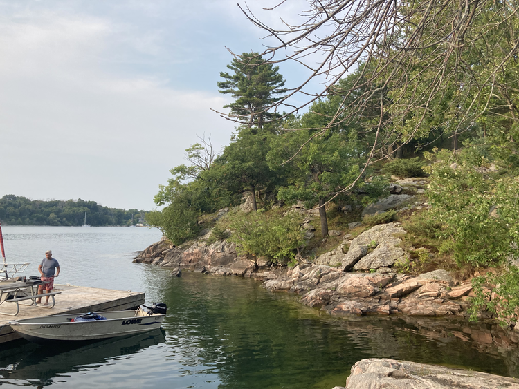 Photo taken by Cindy Ci  Alt text: Landscape shot of the edge of Cedar Island. On the right side is the island, consisting of large rocks, land, and green shrubbery. On the left is the dock with a man standing on it.