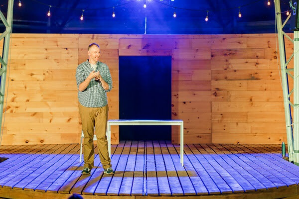 Captions: Crawford stands symmetrically stage right near a white desk and white chair under blue lighting. His elbows rest at his side while he holds his hands near his chest in a counting gesture.
