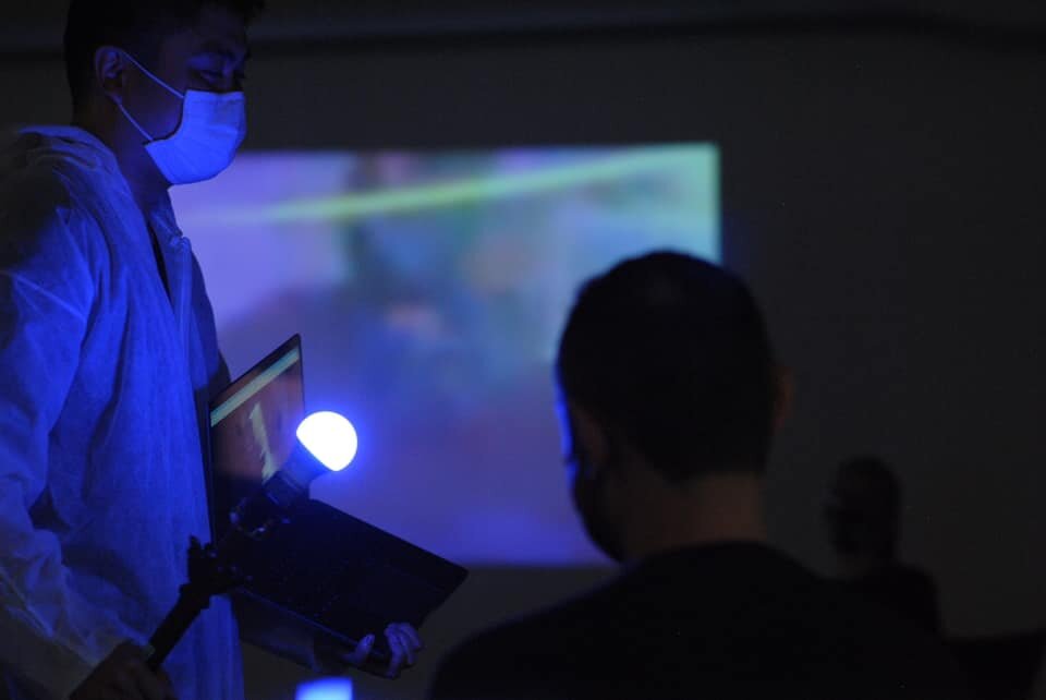 Clayton Lee walks through the audience holding a lightbulb on a stick, and a laptop on a video call with Michael Rubenfeld in Krakow who is doing the same. Both live feeds are projected on the wall.