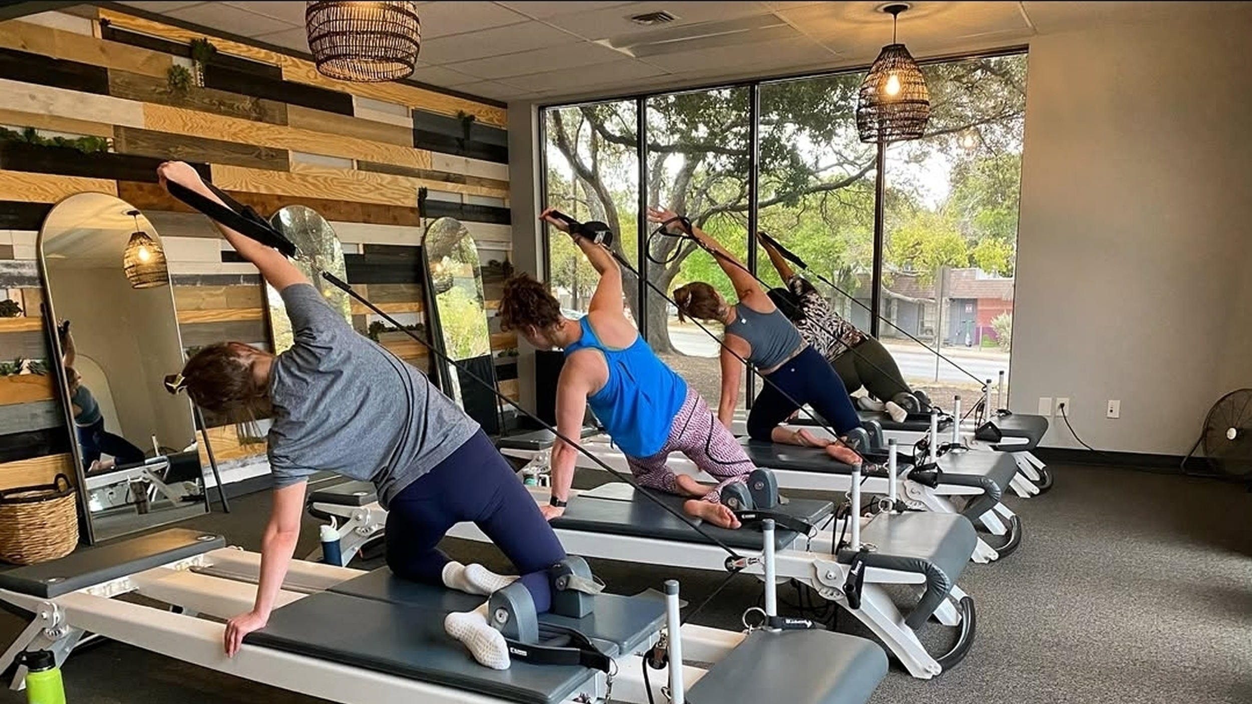 Four women in a Pilates or yoga studio performing side stretches on reformer machines. The studio has wooden wall decor with mirrors, large windows, and hanging woven light fixtures.