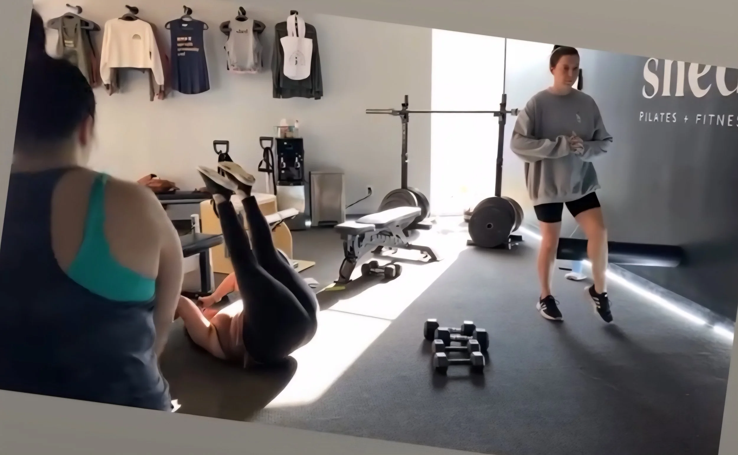 People working out in a gym with exercise equipment, weights, and fitness gear visible.