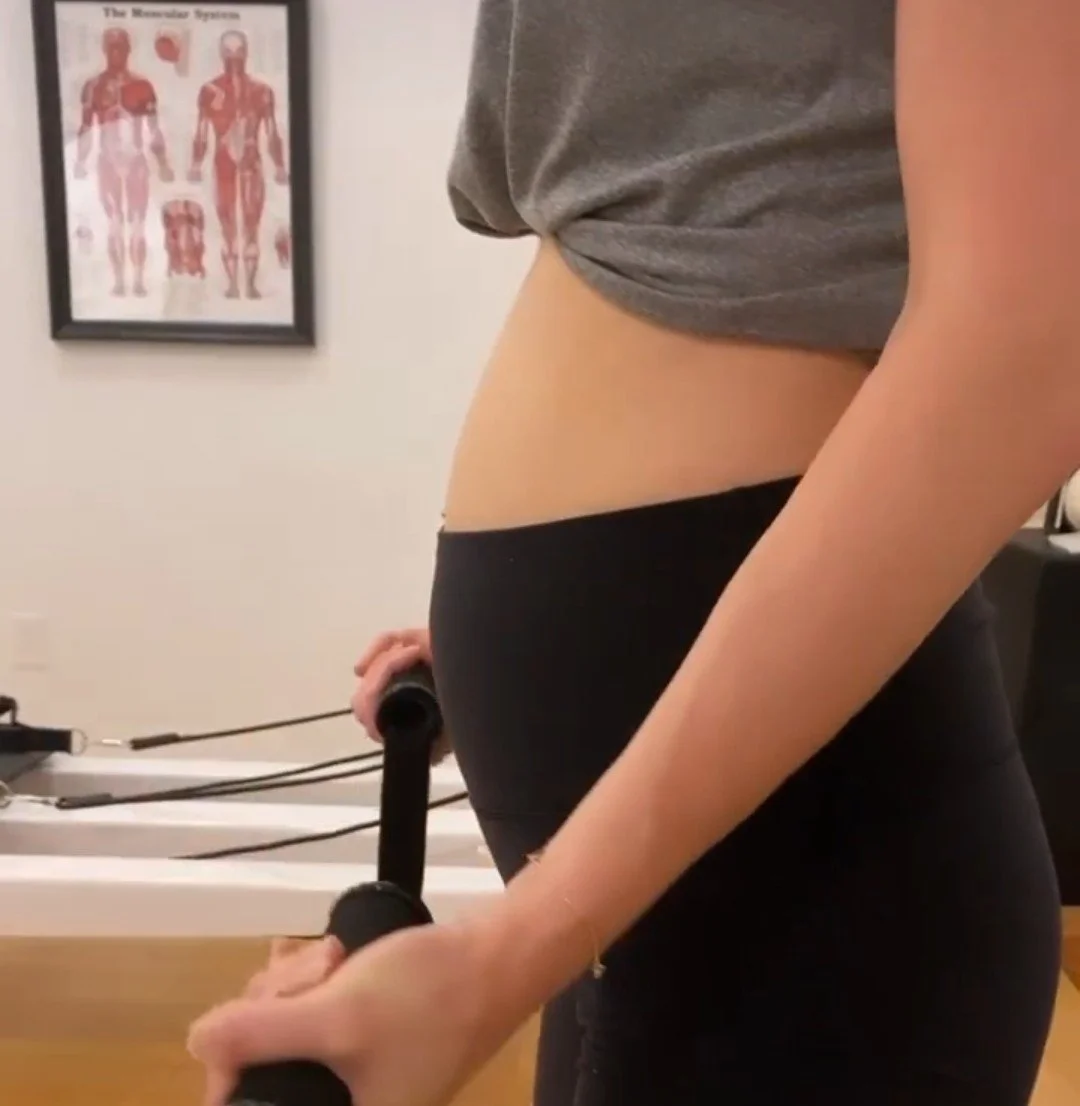 Close-up of a pregnant woman in workout clothes holding handles of a reformer machine, with a medical poster of the human muscular system in the background.