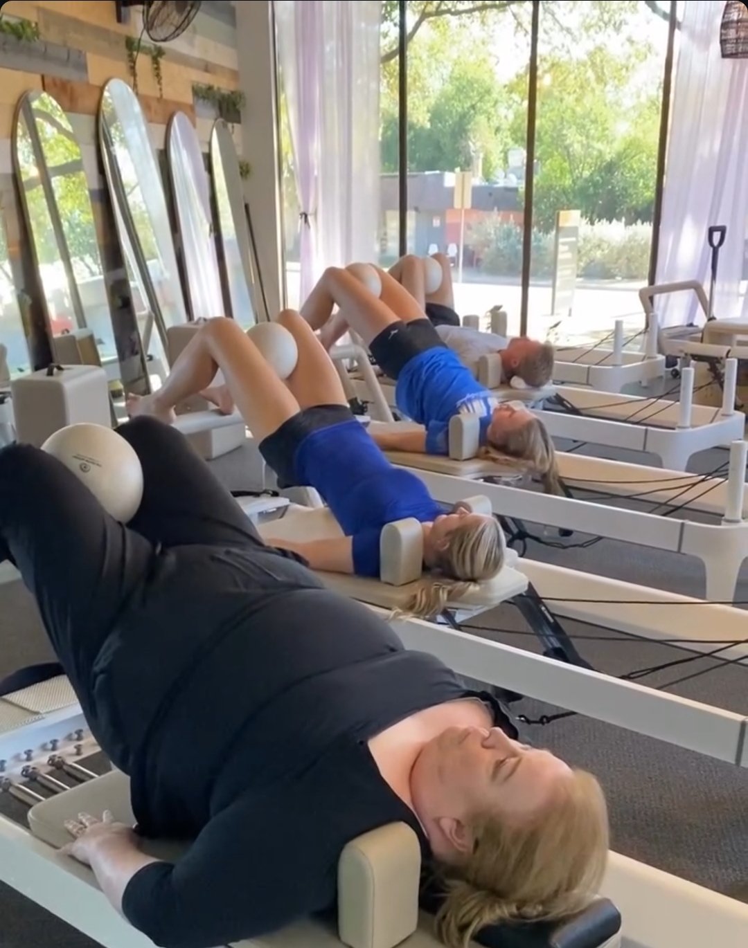 Women doing a bridge and exercising on Pilates reformer machines, with a window in the background.