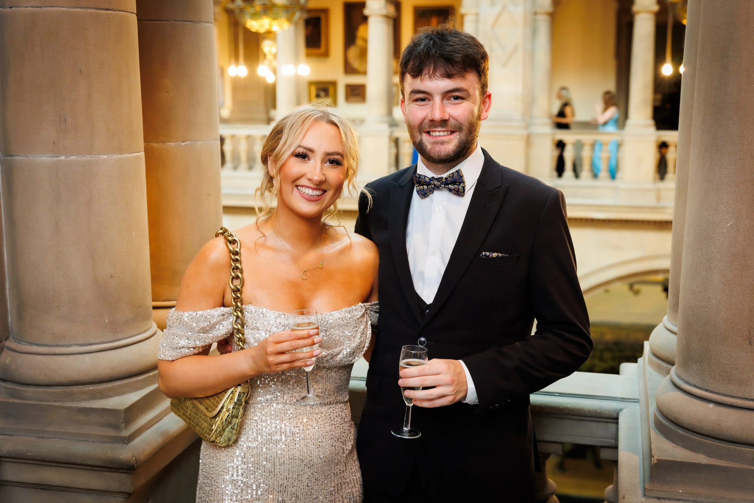 Two guests smiling with champagne in Kelvingrove Art Gallery during a formal event.