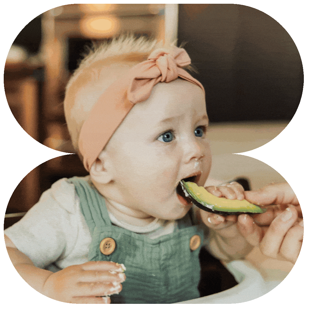 A young child with blonde hair, wearing a peach headband and green overalls, is being fed a slice of avocado.