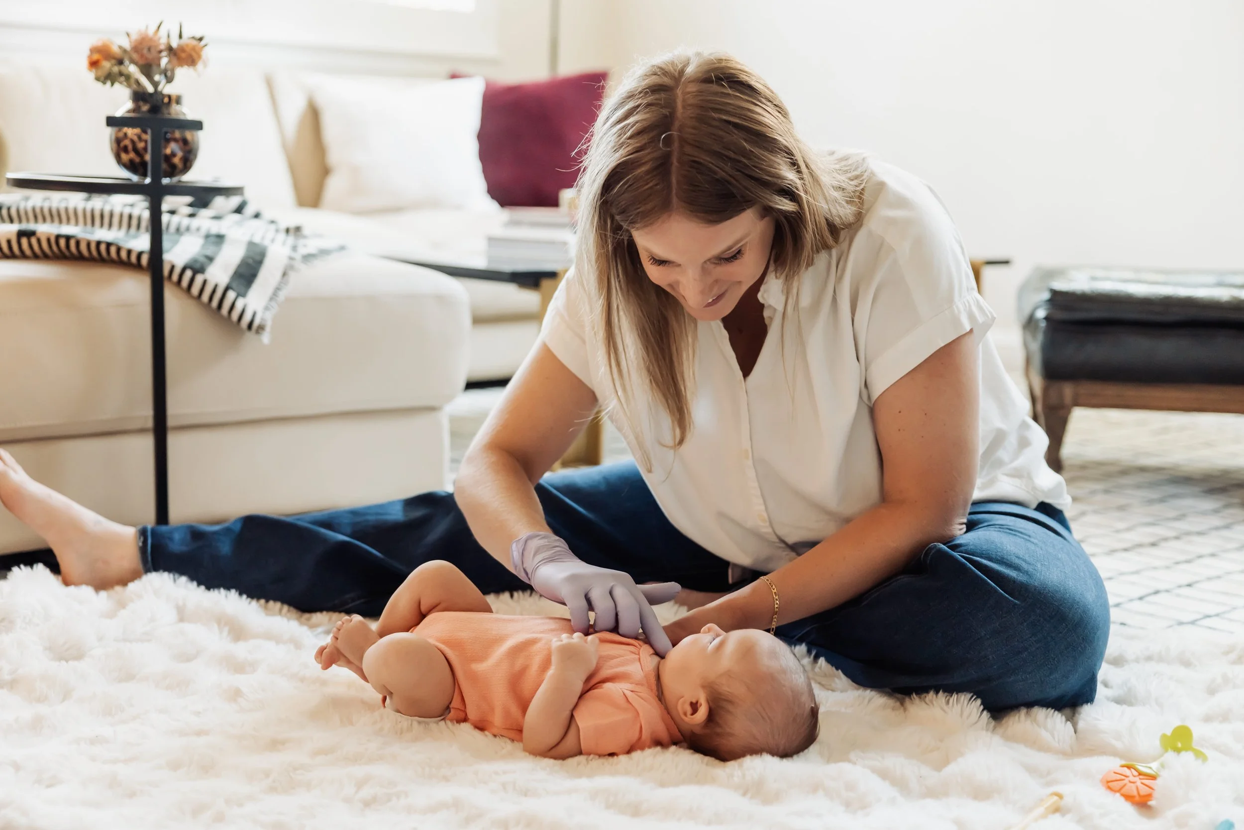 A woman in white shirt and jeans performing infant CPR on a baby lying on a plush white rug in a living room.