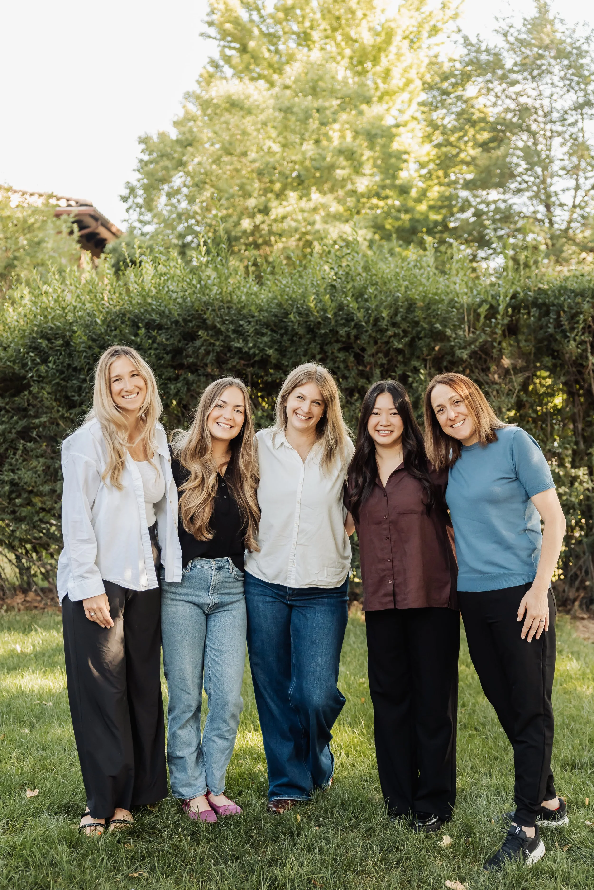 Group of five women standing outdoors on grass, smiling, with trees and sunlight in the background.