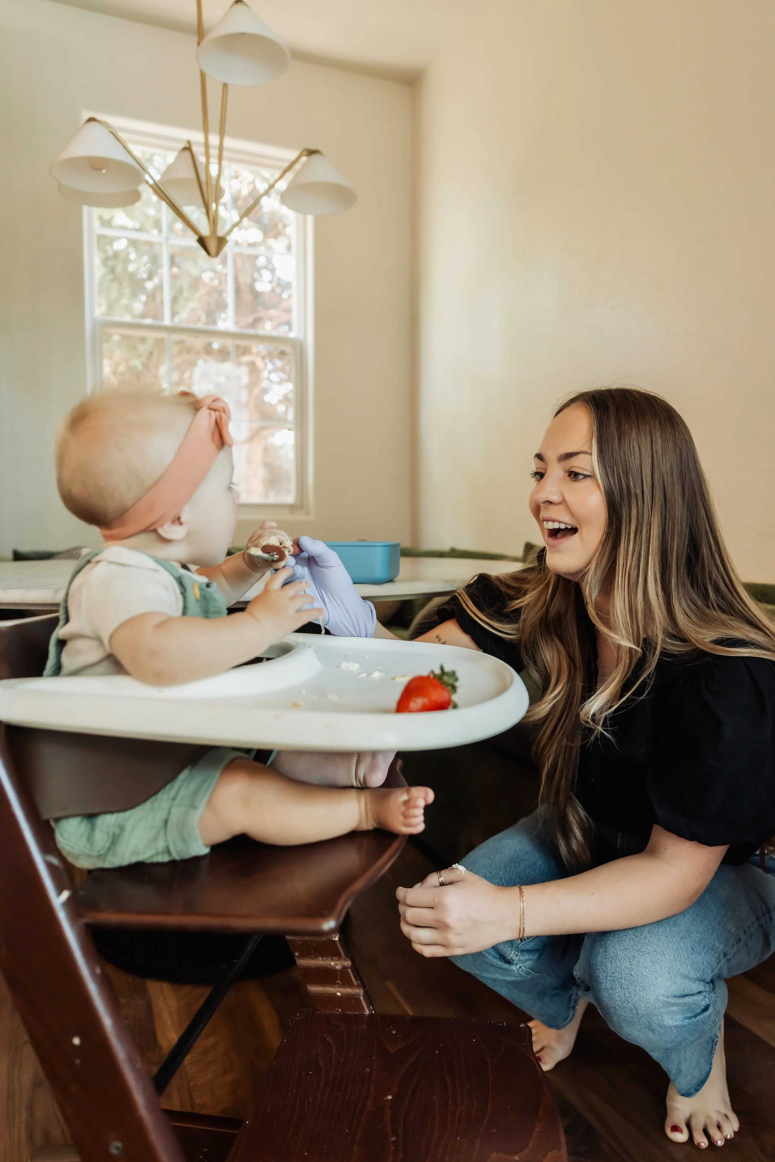 A woman with long brown hair, wearing blue jeans and a black top, kneeling on the floor and playing with a toddler in a high chair. The toddler has blonde hair and is wearing a white shirt with suspenders, holding a spoon. There is a strawberry on the high chair tray. The room has a window in the background and a chandelier hanging from the ceiling.