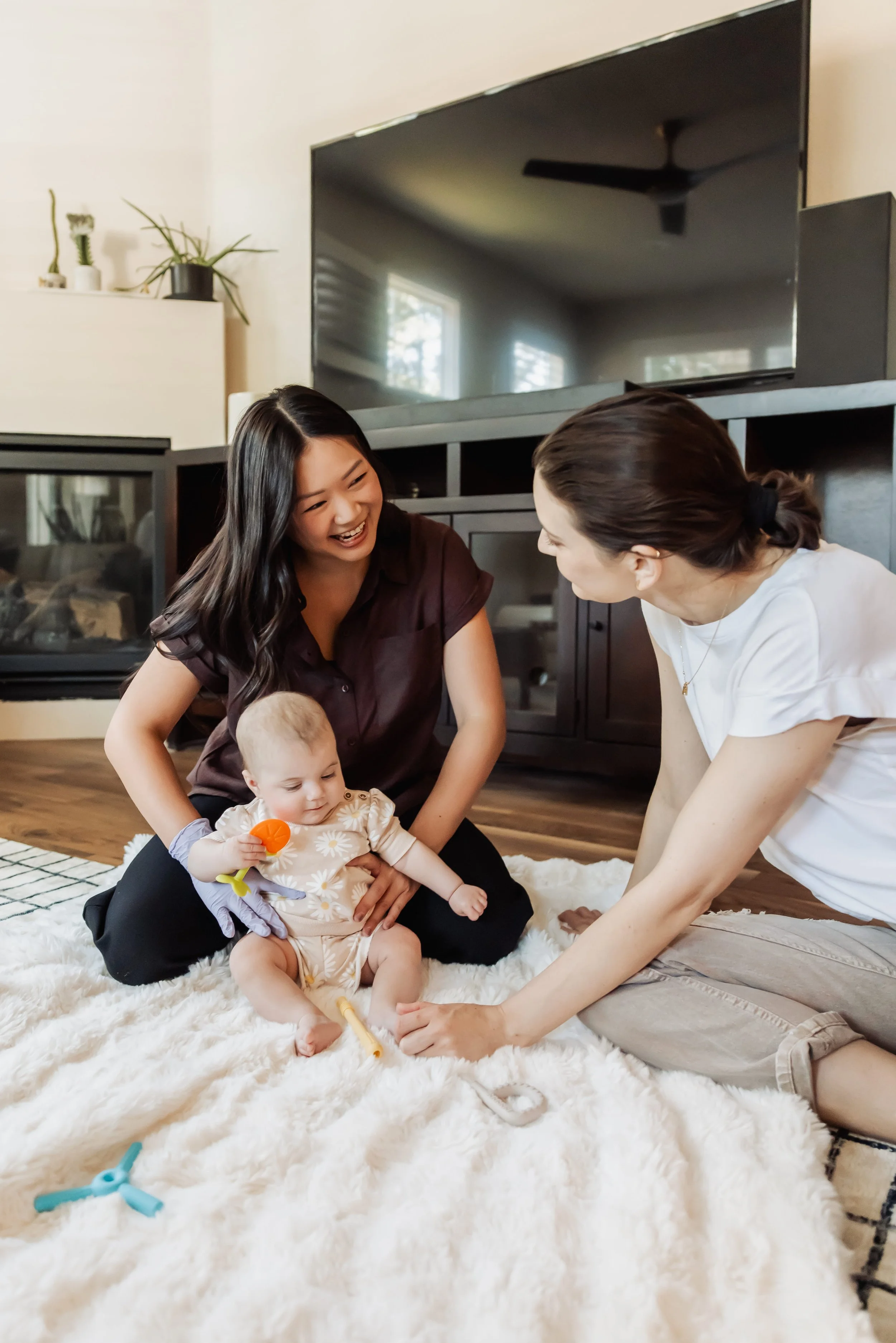 Two women, one with black hair and the other with brown hair, sitting on a white fluffy rug, playing with a baby on a cozy living room floor. The woman with black hair is smiling, while the other woman leans in and holds the baby's hand. The baby, wearing a beige outfit with a daisy pattern, is holding an orange toy and a yellow medical instrument. In the background, there is a black cabinet, a large flat-screen TV, and some potted plants.