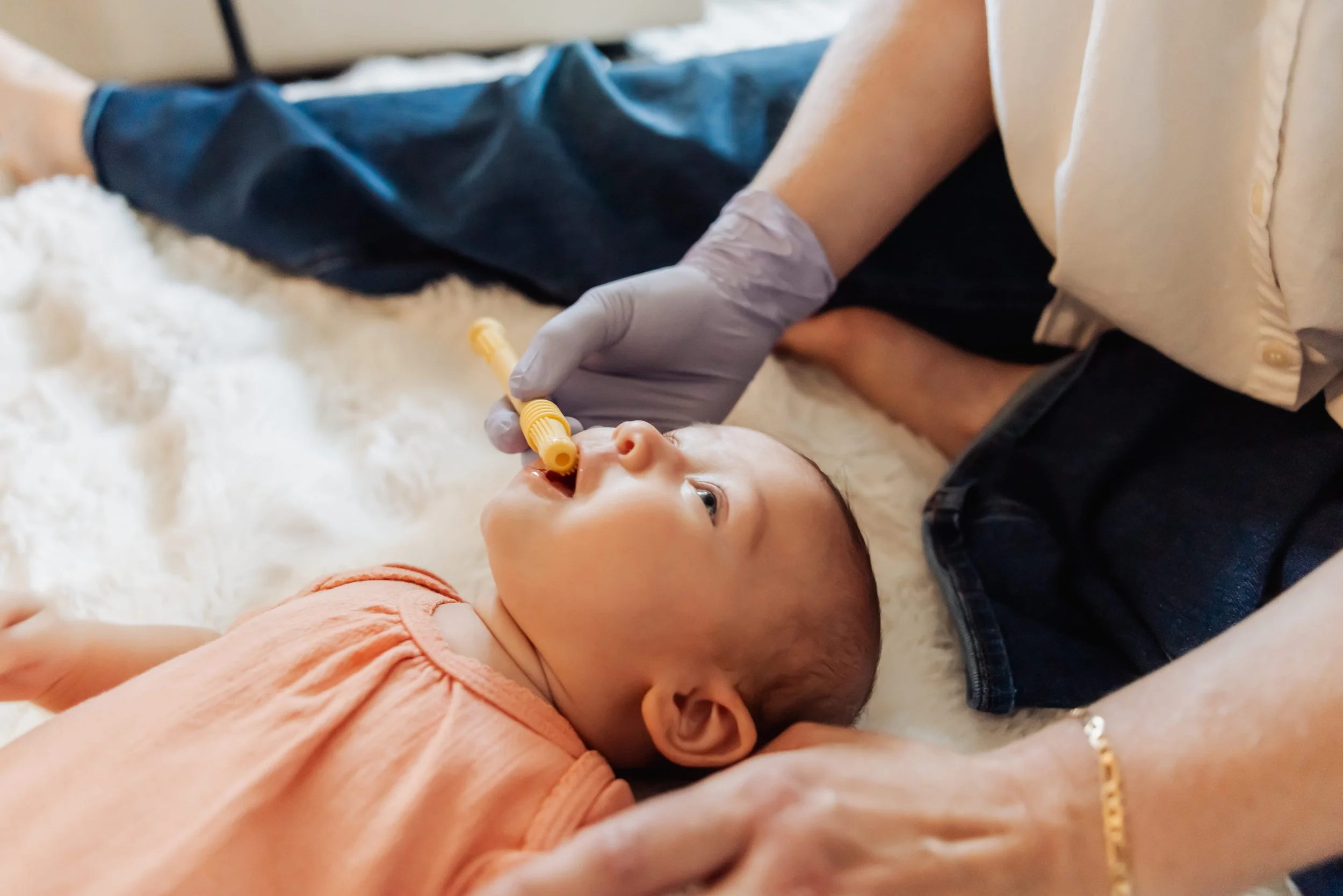 A young child lying on a soft surface with a caregiver near, holding a yellow nasal aspirator to the child's nose.