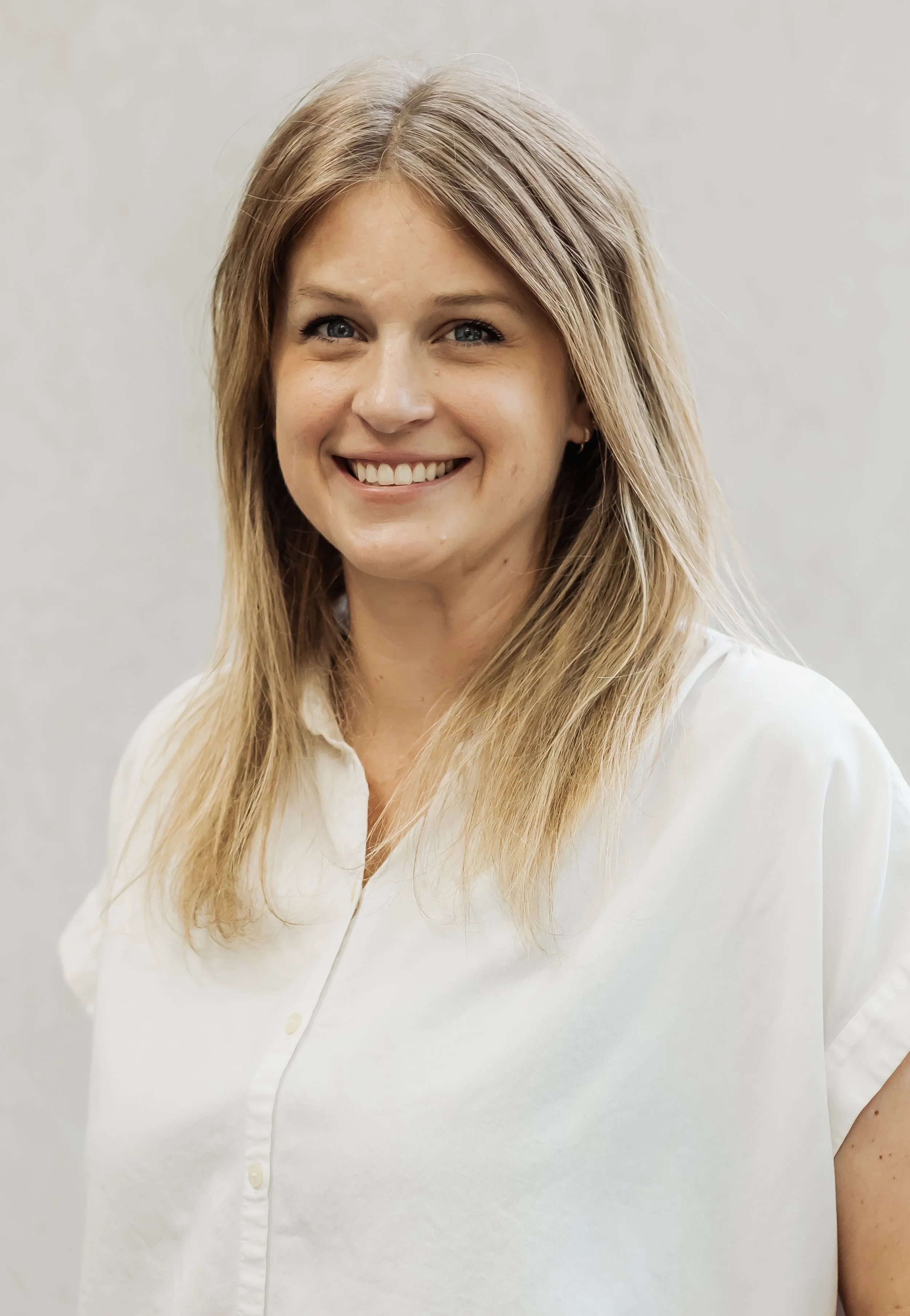 Close-up of a woman with blonde hair wearing a white shirt, smiling, against a plain light-colored background.