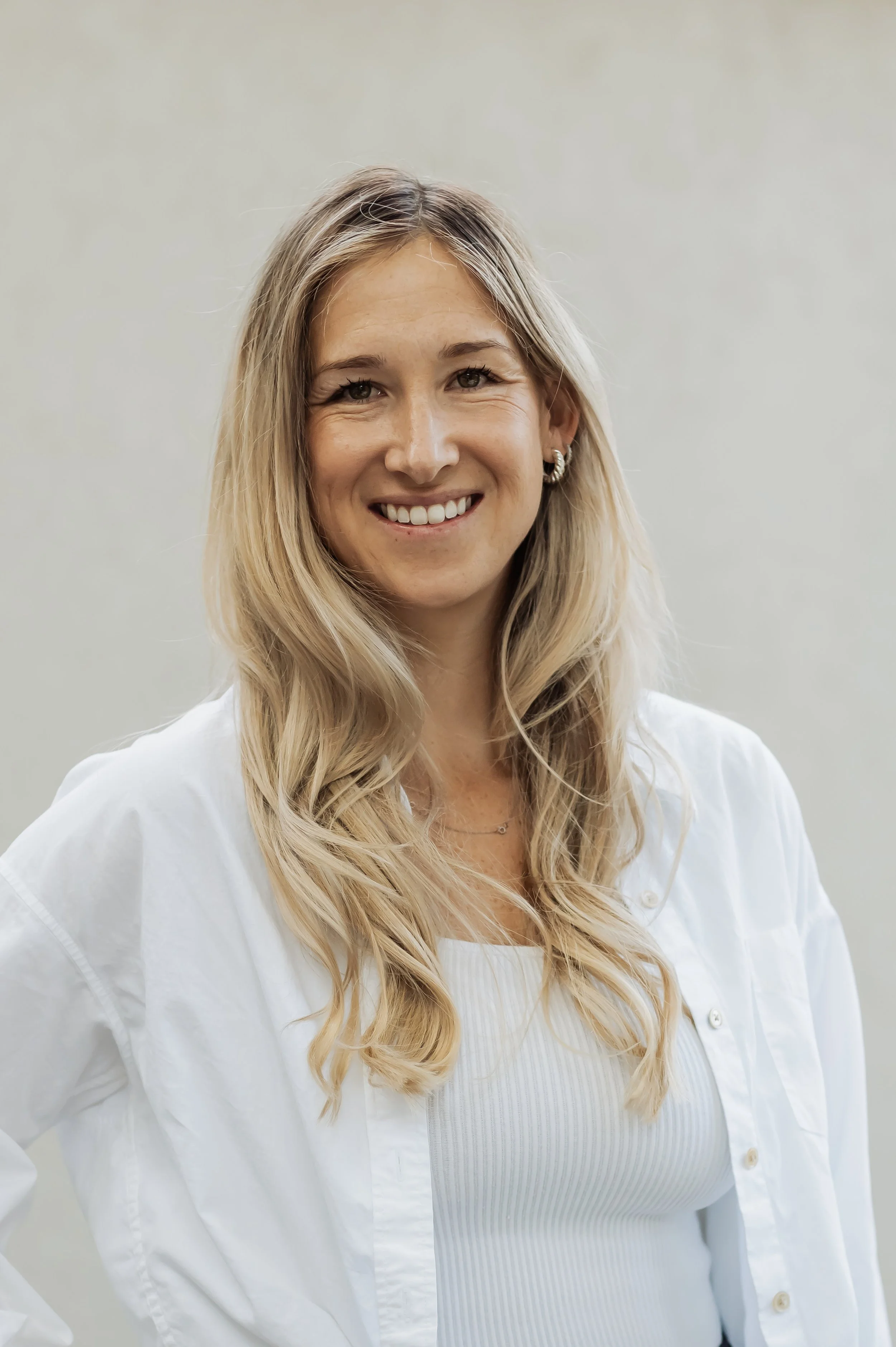 A woman with long wavy blonde hair smiling at the camera, wearing a white button-up shirt and silver hoop earrings, standing against a plain light-colored background.