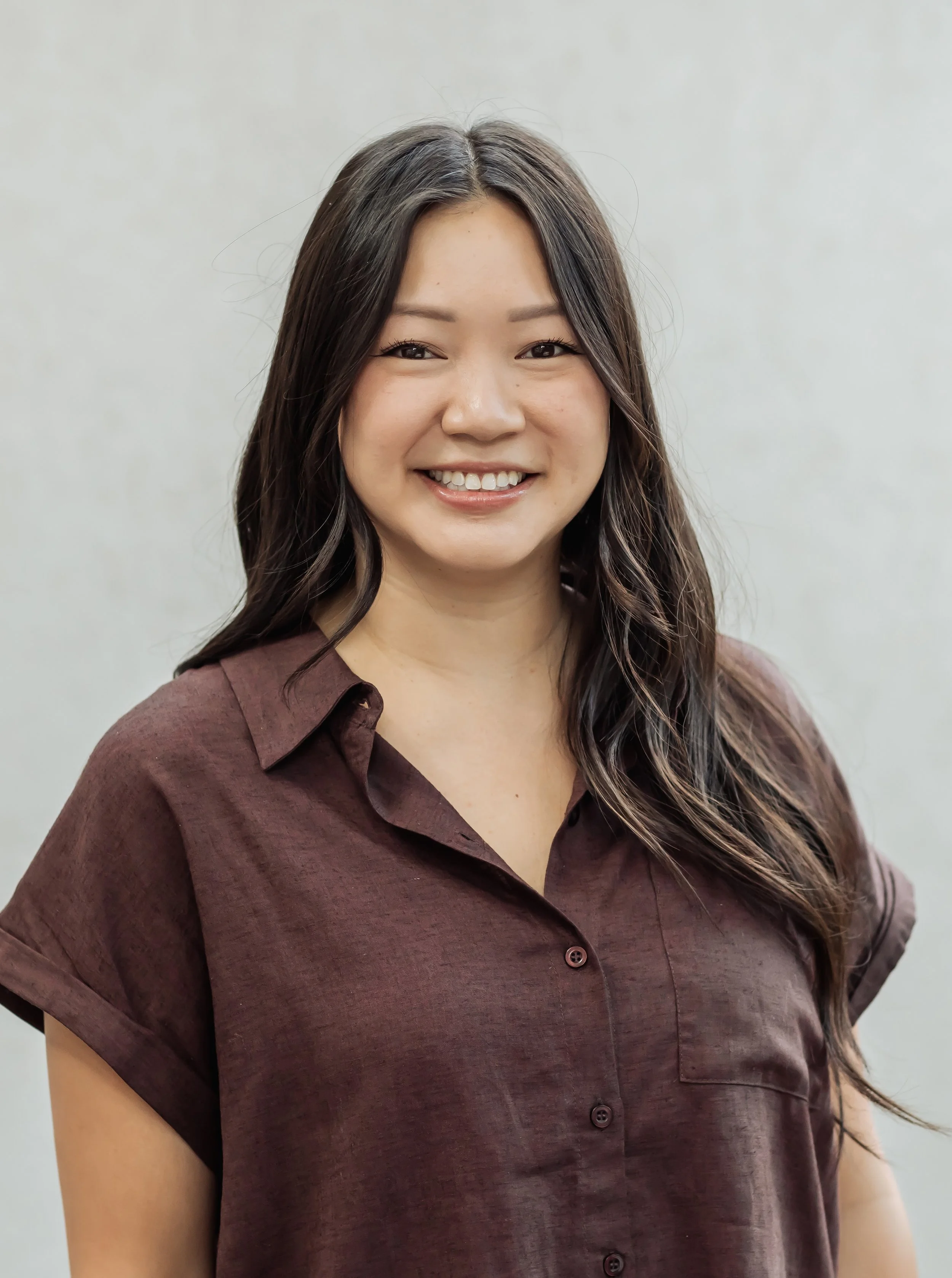 Close-up of a smiling woman with long, wavy dark hair wearing a brown button-up shirt against a plain light background.