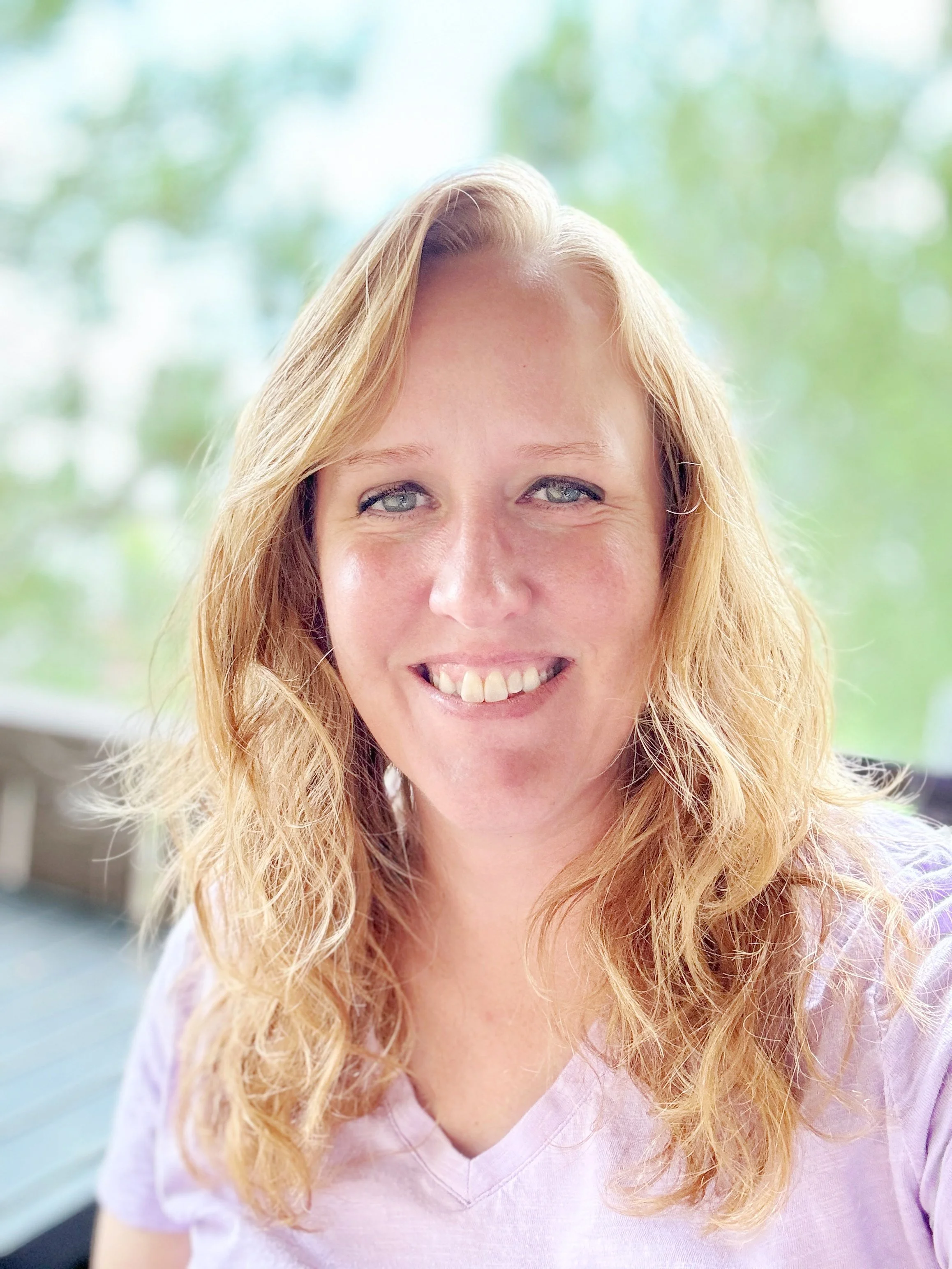 Close-up of a smiling woman with long, wavy blonde hair and blue eyes, wearing a light pink top, with blurred green trees in the background.