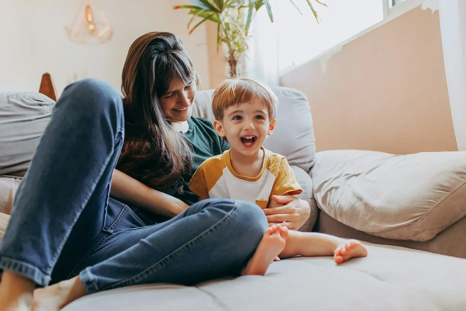Excited young boy sits with his mom on a couch where both are smiling.