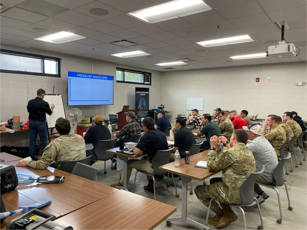 A classroom setting with people taking notes and listening to a instructor at the front. The instructor is writing on a whiteboard, and a large screen displays a presentation slide titled 'Pressure Waveform.' The room has beige walls, ceiling lights, and windows near the ceiling.