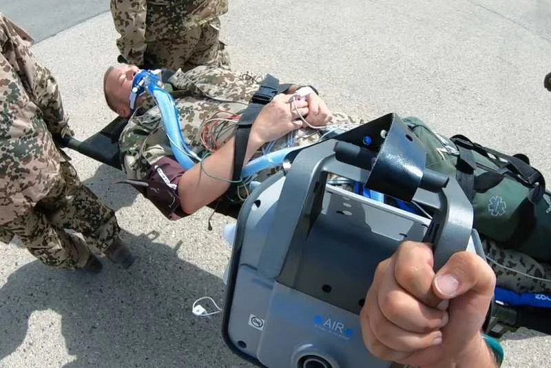 Person being evacuated on a stretcher with medical equipment, surrounded by military personnel.