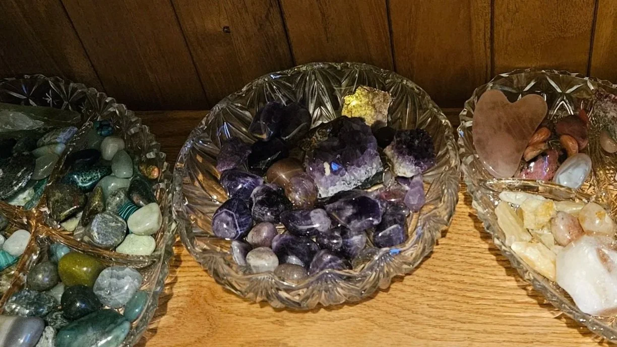 three bowls of crystals sit next to each other on a shelf. The one on the right hand side is long and contains various shades of green, the middle one is smaller and rounder and contains purples, and the one of the left is longer, with white and pink