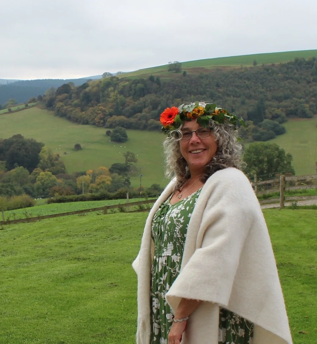 A white woman with grey curly hair and black glasses stands in front of some fields, she is wearing a green pattern dress and a white shawl. There is a garland of orange and yellow flowers in her hair.
