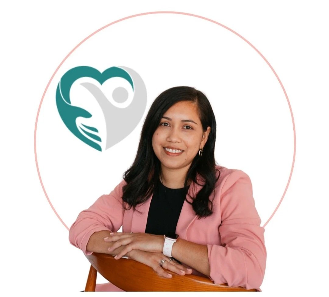 A woman with black hair and a pink blazer sitting with her arms crossed on a wooden chair, smiling at the camera with a logo of a heart-shaped design with a person inside and two hands forming the heart behind her on a white background.