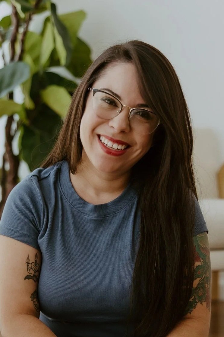 A smiling woman with long dark hair, glasses, and tattoos on her arms, sitting indoors with a plant in the background.