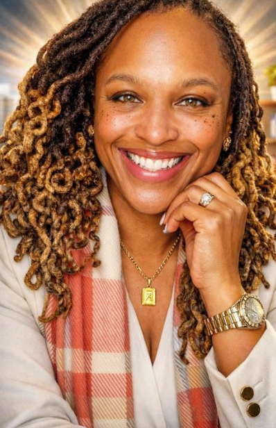 Portrait of a smiling woman with curly, ombré hair, wearing a white blazer with a pink plaid scarf, gold watch, necklace, and ring.