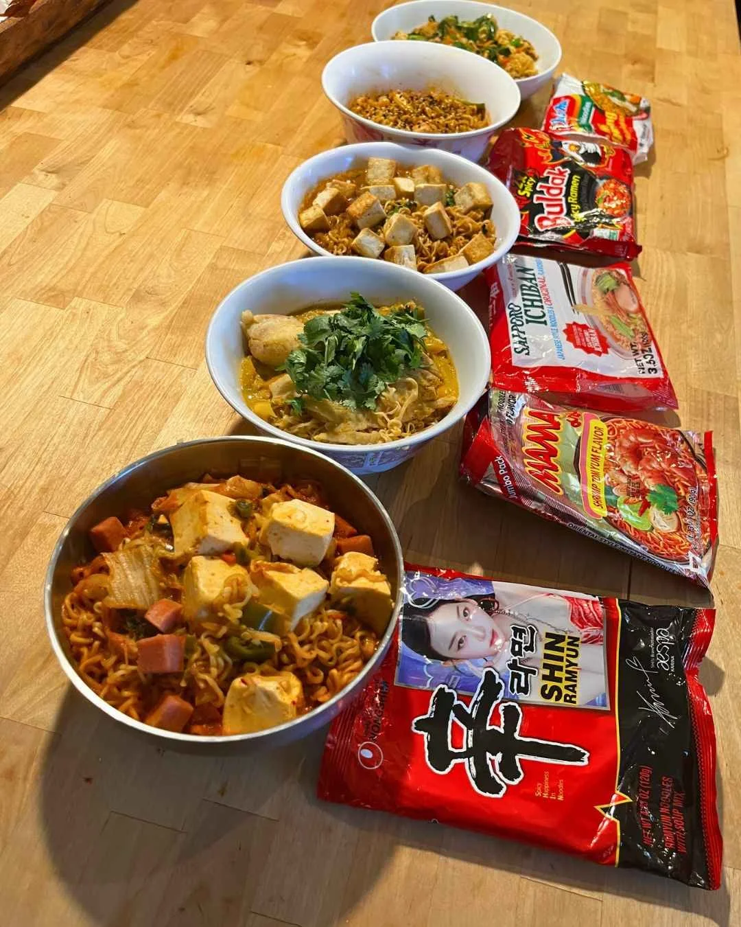 Five finished noodle dishes from Instant Noodles and Beyond, each next to its instant noodle brand package