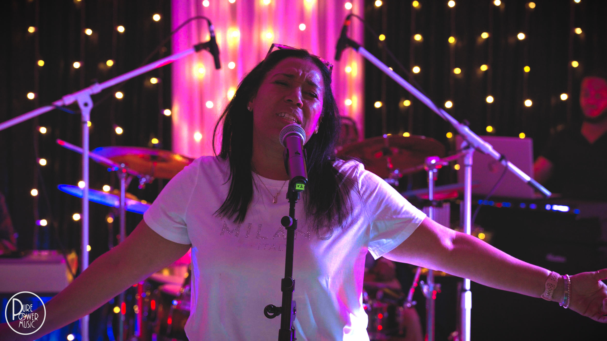 Singer performing on stage with a microphone in front of drums and colorful stage lighting.