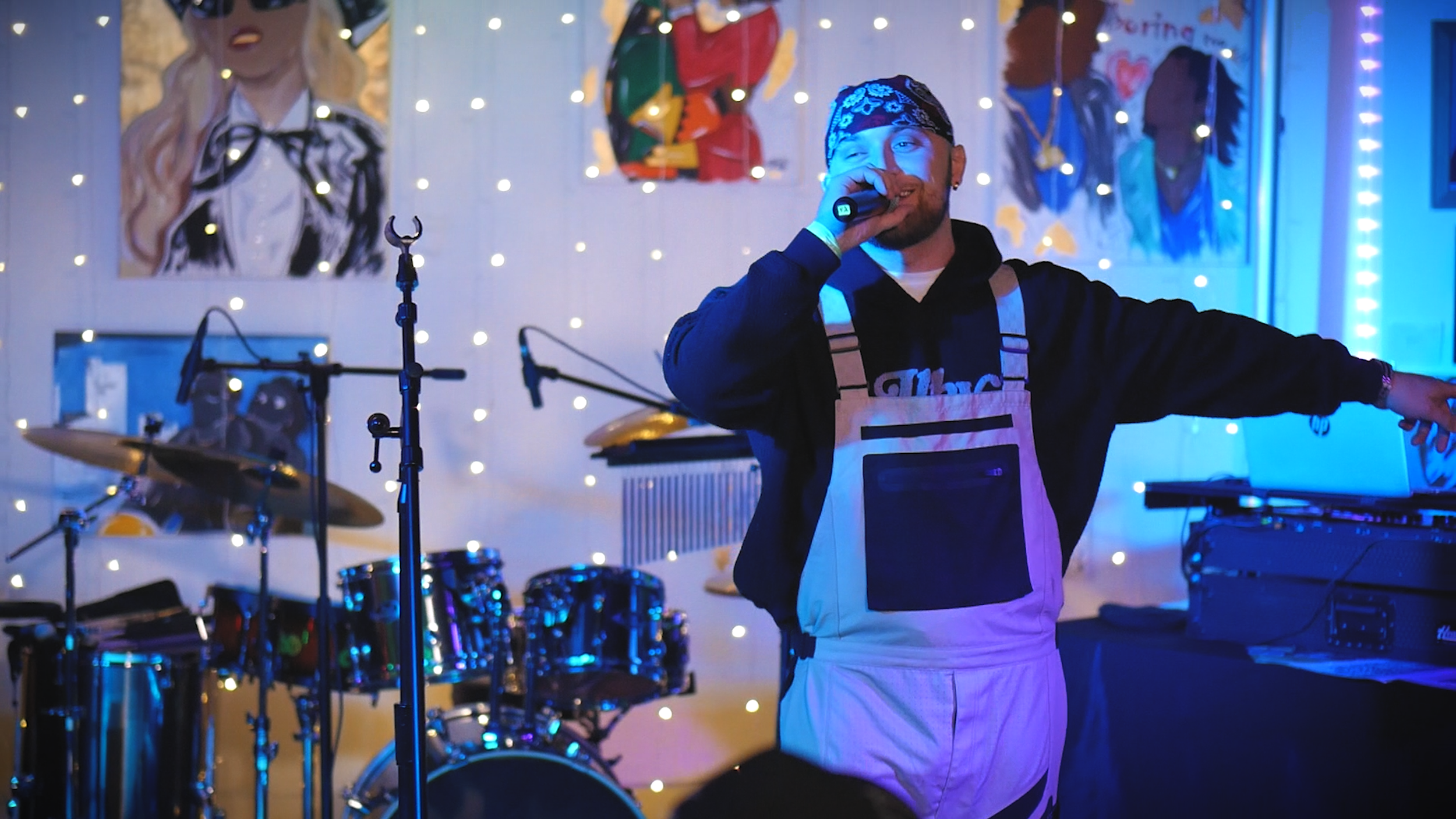 Vocalist performing on stage with a microphone in front of a drum kit and colorful studio lighting.