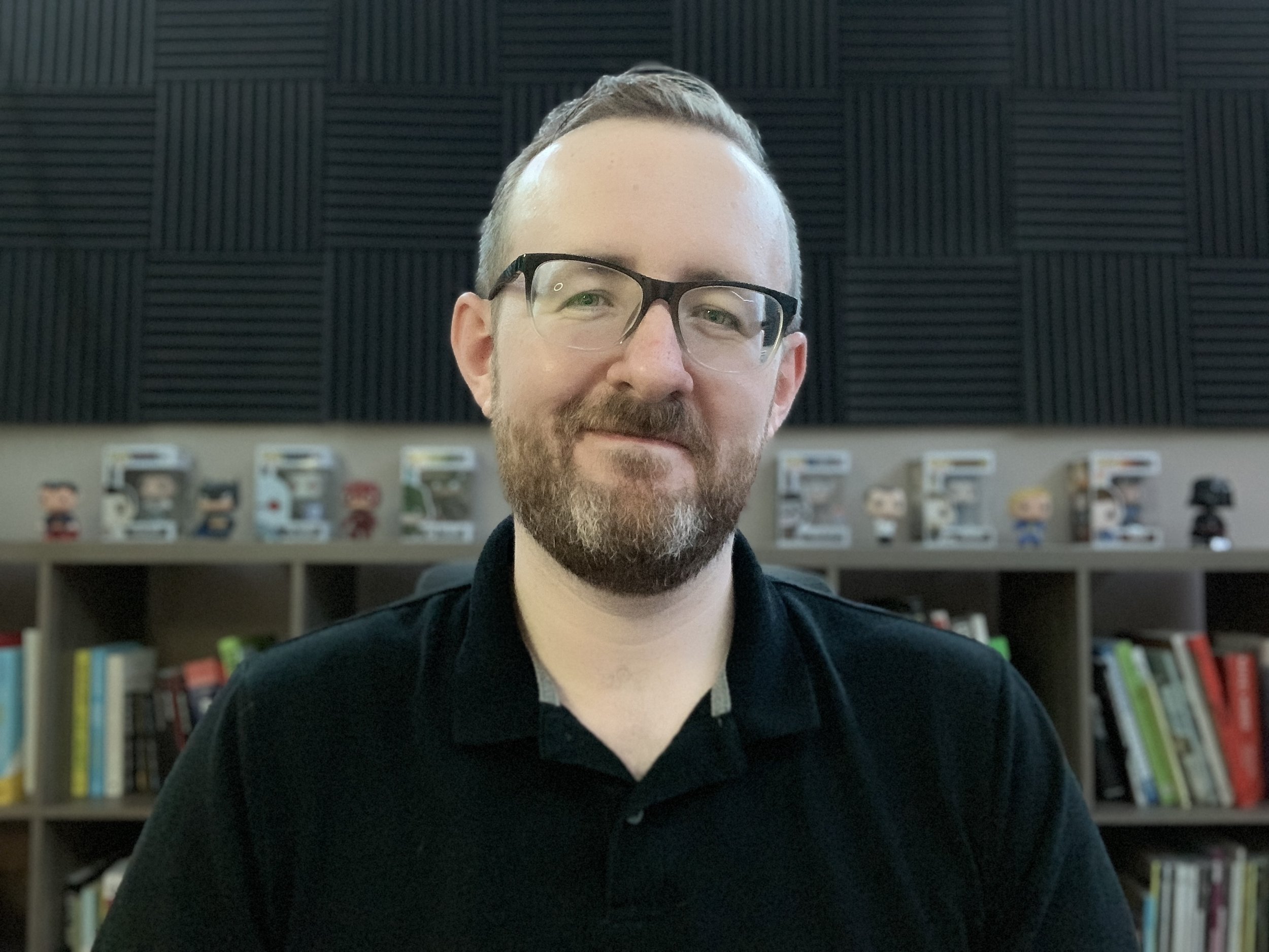 A man with glasses and a beard, wearing a black shirt, sitting in front of a bookshelf with books and Funko Pop figures.
