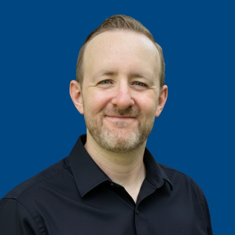 A man with short hair and a beard, wearing a black collared shirt, smiling at the camera against a blue background.