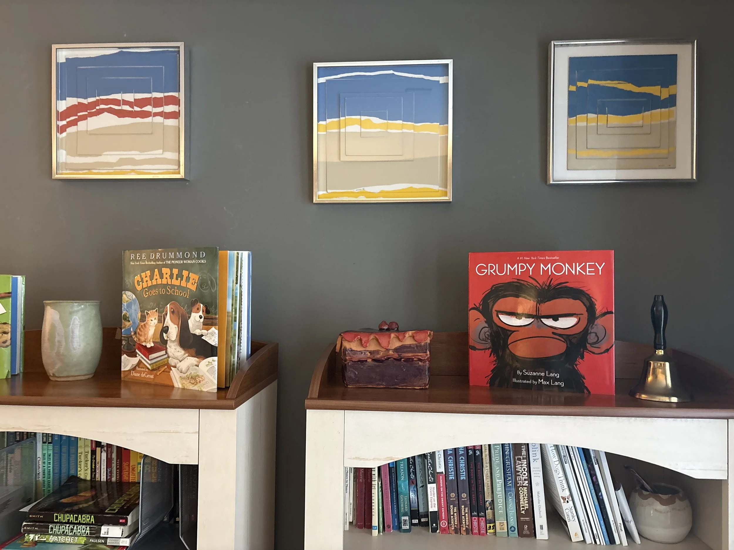 Books and framed artwork arranged on built-in shelves against a gray wall. The top shelf features three framed abstract landscape paintings with blue, beige, white, red, and yellow colors. The middle shelf displays children's books, including titles 