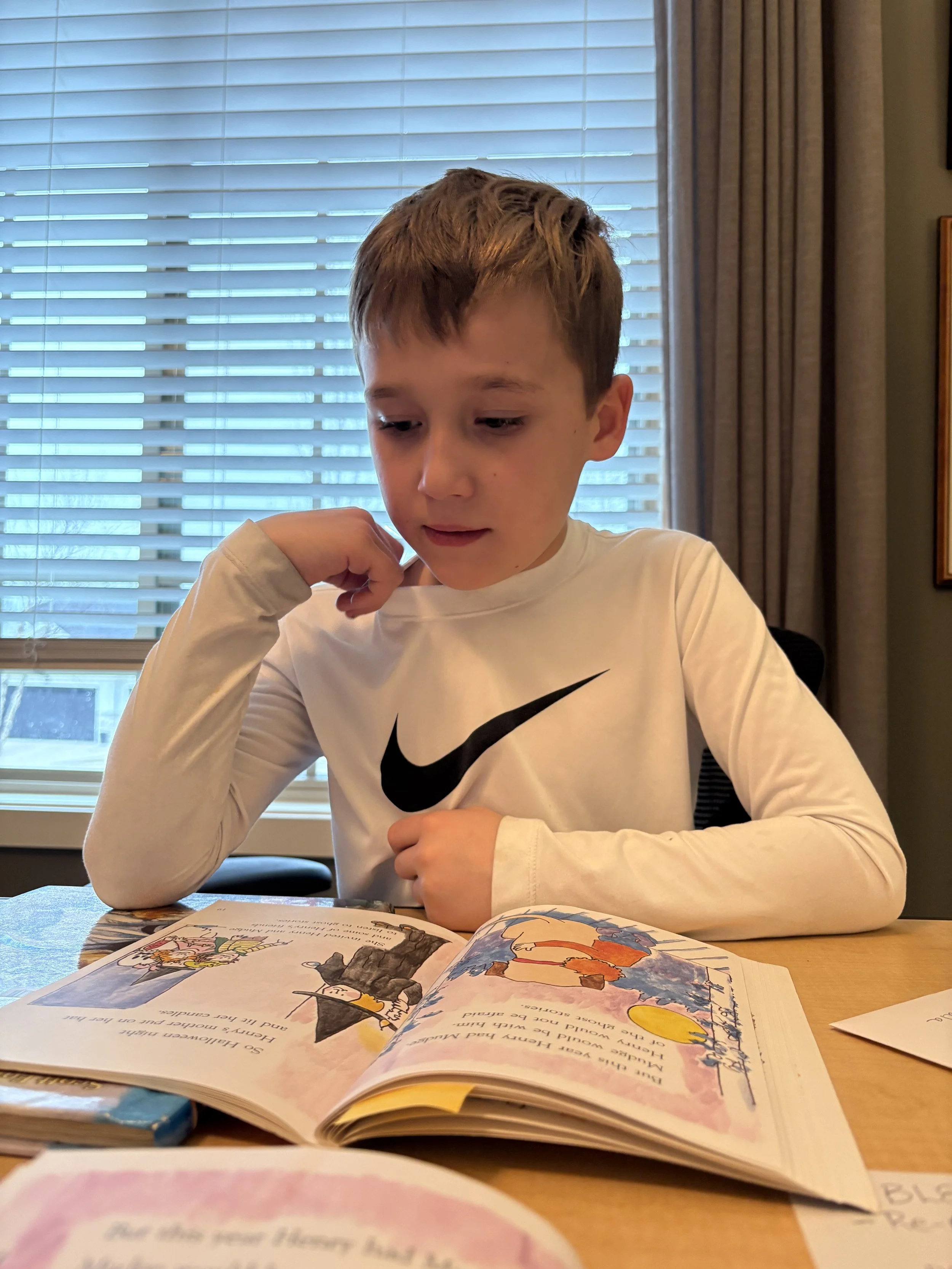 A young boy with short, light brown hair, wearing a white long-sleeved shirt with a black Nike logo, is sitting at a table reading a children's book. The open book features colorful illustrations and text. The background includes a window with blinds