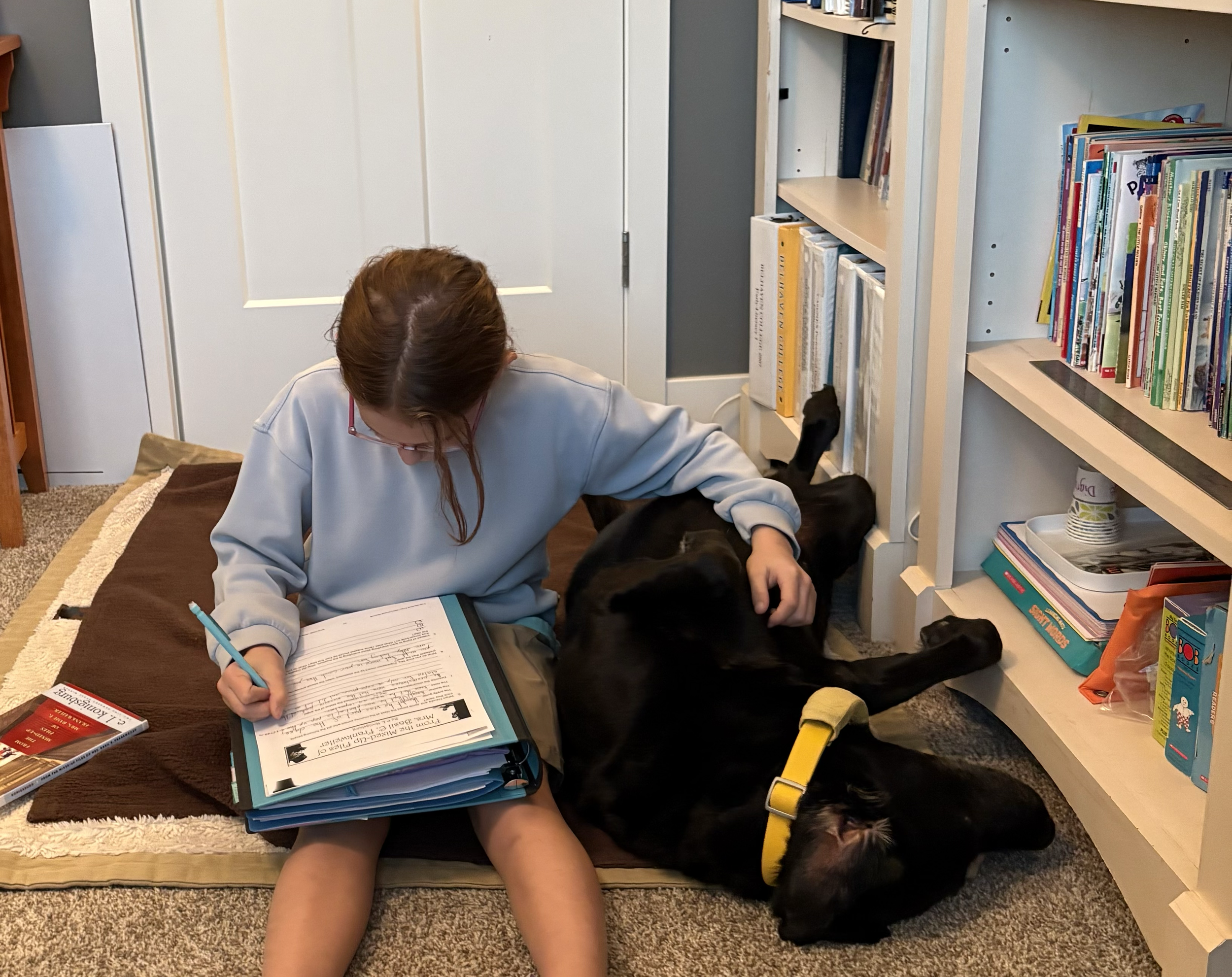 A person with brown hair wearing glasses is sitting on the floor in a room, reading and writing in a workbook, with a black dog wearing a yellow collar lying beside them. Bookshelves with colorful books are behind them.